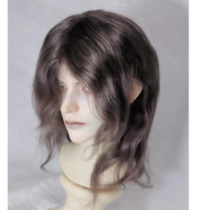 May include: A doll head with a wig. The wig is a wavy, dark gray color. The doll has pale skin, a neutral expression, and is mounted on a cream-colored stand. The doll's features are finely detailed, with a focus on the eyes and lips.