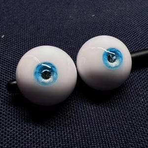 Op de afbeelding: Twee witte poppenogen met blauwe iris en zwarte pupillen. De ogen zijn licht glanzend en hebben een realistisch uiterlijk.