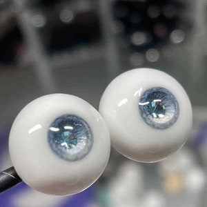 Op de afbeelding: Twee witte glazen poppenogen met blauwe iris en een glinsterend, lichtblauw centrum. De ogen staan op een zwarte standaard.