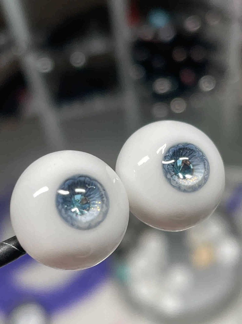 Op de afbeelding: Twee witte glazen poppenogen met blauwe iris en een glinsterend, lichtblauw centrum. De ogen staan op een zwarte standaard.
