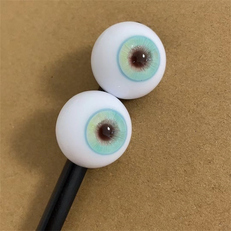 Puede incluir: Dos ojos de mu&ntilde;eca blancos con iris azules y verdes y pupilas marrones. Los ojos est&aacute;n en un palo negro.