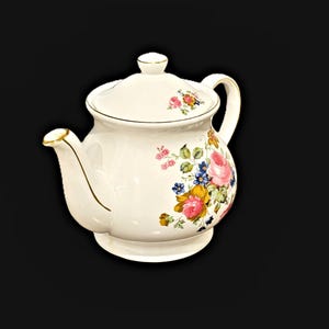 May include: A white ceramic teapot featuring a floral pattern with pink, yellow, and blue flowers and green leaves. The teapot has a matching lid with a small knob, a curved handle, and a spout, all accented with thin gold trim along the edges.
