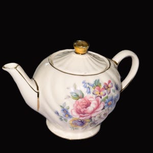 May include: Cream porcelain teapot with a floral pattern of pink roses and blue and yellow flowers. The teapot has gold detailing on the handle, spout, and lid. The lid is topped with a gold knob.