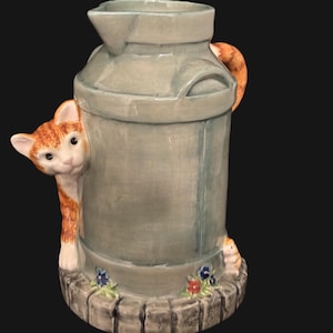 May include: A ceramic milk jug pitcher with a cat peeking out from behind it. The jug is painted to look like a metal milk jug with a handle. The base of the jug is decorated with flowers and a small mouse.