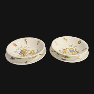May include: Two sets of cream-coloured ceramic bowls and matching plates, each featuring colourful illustrations of teddy bears in blue and red clothing, surrounded by small garden motifs like carrots, potted flowers, and watering cans.