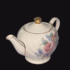 May include: Cream-coloured porcelain teapot with a gold lid and handle. The teapot has a floral design with pink, blue, and purple flowers. The spout and rim are trimmed with gold.