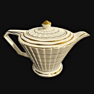 Peut inclure: Une théière en céramique blanche dotée d'un corps cannelé et nervuré, ainsi qu'un couvercle assorti avec un bouton doré. La théière est ornée de bandes dorées horizontales sur le corps et le couvercle, avec une base et une anse bordées d'or.