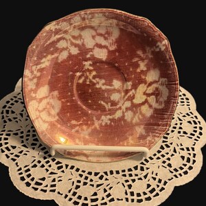 Peut inclure: Une soucoupe en céramique ronde présentant un motif floral rouge foncé et blanc avec une texture en forme de grille. La soucoupe possède un bord festonné et une indentation circulaire centrale pour une tasse, posée sur un napperon en dentelle.