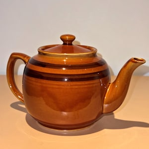 May include: A round ceramic teapot with a glossy finish in shades of amber and brown. The teapot features a matching lid with a small knob, a curved handle on the left, and a tapered spout on the right. Darker brown horizontal bands encircle the body.