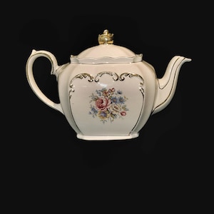 May include: A cream-coloured porcelain teapot with a floral design. The teapot has a curved handle, a spout, and a lid with a gold crown-shaped finial. Gold trim outlines the edges and the floral design, which includes pink, blue, and yellow flowers.
