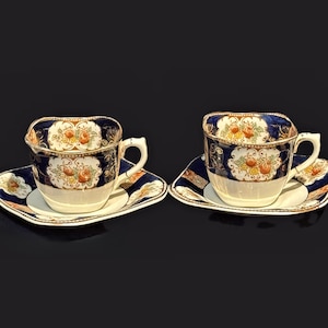 May include: Two antique teacups and saucers. The cups and saucers are white with a navy blue border and floral design. The cups have a unique square shape and gold trim. The saucers are square with a matching design.