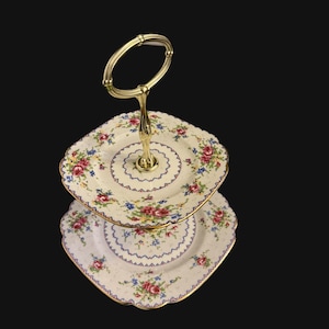 May include: Two-tiered white porcelain cake stand with a gold handle and floral pattern. The stand has a square shape and is decorated with delicate floral designs in shades of pink, blue, and green.