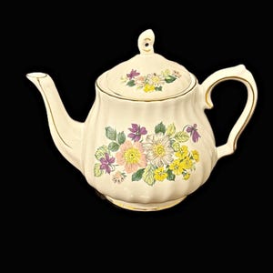 Peut inclure: Une théière en céramique de couleur crème avec un corps côtelé et un couvercle assorti. La théière présente un motif floral coloré avec des fleurs roses, jaunes et violettes et des feuilles vertes. Le bec, l'anse et le bouton sont dorés.