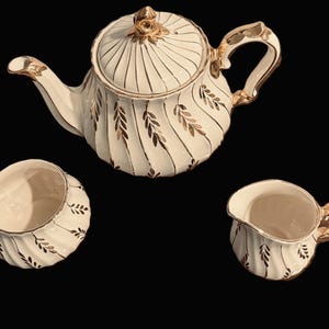 May include: A cream-coloured ceramic tea set consisting of a teapot with a lid, a sugar bowl, and a creamer. Each piece features a vertical ribbed texture decorated with a repeating gold leaf pattern and gold trim on the handles and spouts.