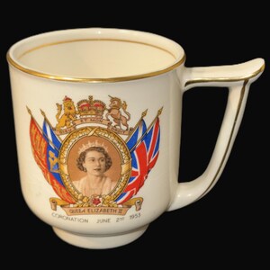 May include: Cream ceramic mug with gold trim, commemorating the coronation of Queen Elizabeth II on June 2nd, 1953. The mug displays a portrait of the Queen, flags, and a royal crest. It has a distinctive handle.