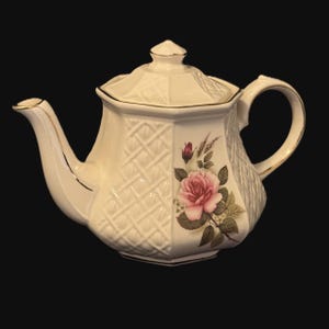 May include: Cream-coloured ceramic teapot with a floral rose design. The teapot has a textured lattice pattern and gold trim. The lid has a small finial, and the handle and spout are curved.