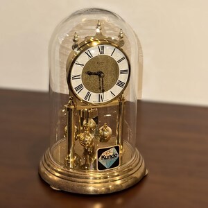 May include: A gold-tone Kundo quartz anniversary clock encased in a clear glass dome. The clock features a white dial with black Roman numerals, a gold centre with a floral pattern, and a rotating pendulum with four gold spheres at the base.