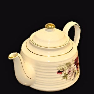 May include: A cream-coloured ceramic teapot featuring horizontal ribbed detailing, a gold-trimmed lid with a gold knob, and a gold-rimmed spout. The body is decorated with a floral pattern of white, pink, and deep red roses with green leaves.
