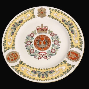May include: A cream-coloured decorative plate with a yellow border. The plate features a central emblem with a stag, surrounded by a floral wreath and a crown. The border includes text and decorative elements.
