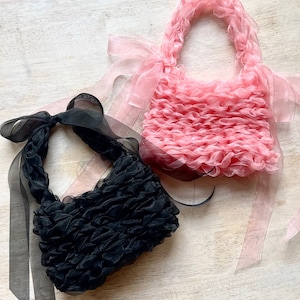 Pode incluir: Duas pequenas bolsas de tecido franzido, uma preta e outra rosa. Ambas têm uma alça trançada e laços longos e transparentes. As bolsas são feitas de um material macio e texturizado, sobre um fundo de madeira clara.