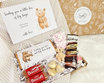 May include: A gift box filled with treats, including a teddy bear, marshmallows, hot chocolate, chocolate hearts, and Biscoff cookies. The box is decorated with a brown and white polka dot pattern and a card that says "Sending you a little box of big hugs!"