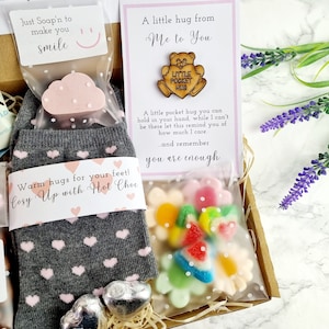 May include: A gift box filled with items for self-care. The box includes a pair of grey socks with pink hearts, a pink cloud-shaped bath bomb, a bag of colourful sweets, and a wooden card with the text "A little hug from Me to You" and a small wooden bear with the text "Little Pocket Hug".