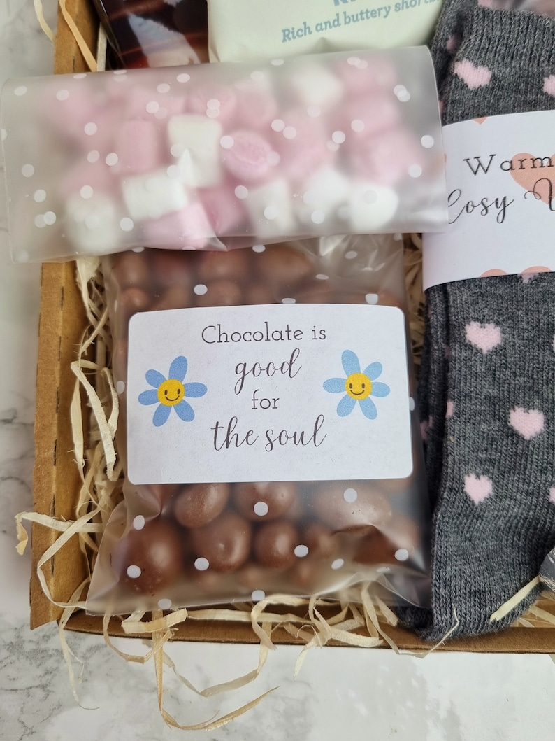 May include: A gift box filled with brown shredded paper, a bag of chocolate sweets, a bag of pink and white marshmallows, and a pair of grey socks with pink hearts. The bag of chocolate sweets has a label that says "Chocolate is good for the soul" with a smiley face flower on each side.