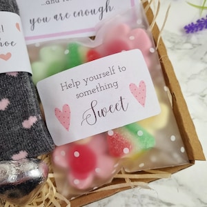 May include: A gift box filled with a grey and pink sock, a silver heart-shaped chocolate, and a clear bag of pink and green gummy sweets. The bag has a white tag with pink hearts and the text "Help yourself to something sweet".