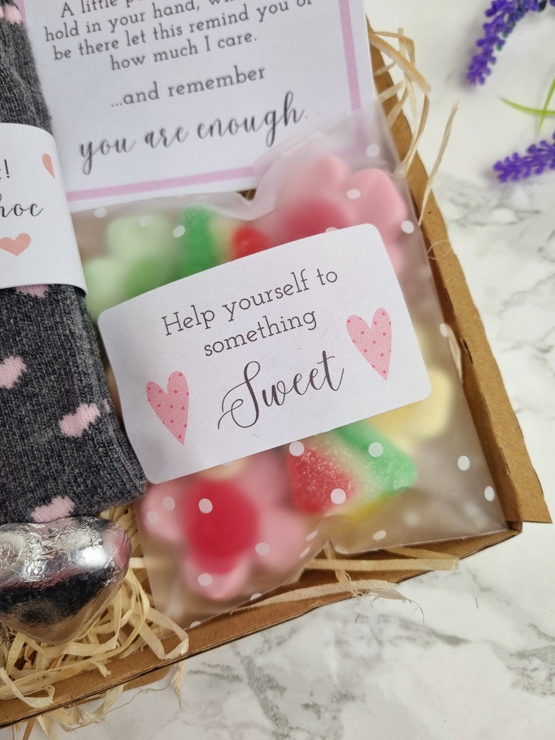 May include: A gift box filled with a grey and pink sock, a silver heart-shaped chocolate, and a clear bag of pink and green gummy sweets. The bag has a white tag with pink hearts and the text "Help yourself to something sweet".