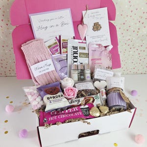 May include: A pink gift box, a 'Hug in a Box', filled with treats. Contains socks, tea, cookies, a candle, and a face mask. Decorated with cards, a heart ornament, and confetti. A thoughtful gift.