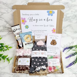 May include: A brown cardboard box filled with a variety of gifts, including a pair of grey socks with pink hearts, a small bar of soap, a bag of chocolate sweets, a bag of marshmallows, a bag of biscuits, a small wooden bear figurine, and a card that says "A little hug from Me to You". The box is decorated with a tag that says "Sending you a little Hug in a box to share some Good Vibes".