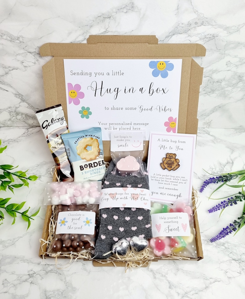 May include: A brown cardboard box filled with a variety of gifts, including a pair of grey socks with pink hearts, a small bar of soap, a bag of chocolate sweets, a bag of marshmallows, a bag of biscuits, a small wooden bear figurine, and a card that says "A little hug from Me to You". The box is decorated with a tag that says "Sending you a little Hug in a box to share some Good Vibes".