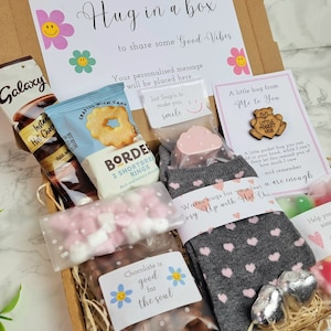 May include: A gift box filled with various items, including chocolate, hot chocolate, socks, and a small wooden sign that says "A little hug from me to you". The box is decorated with a sign that says "Sending you a little hug in a box to share some good vibes".