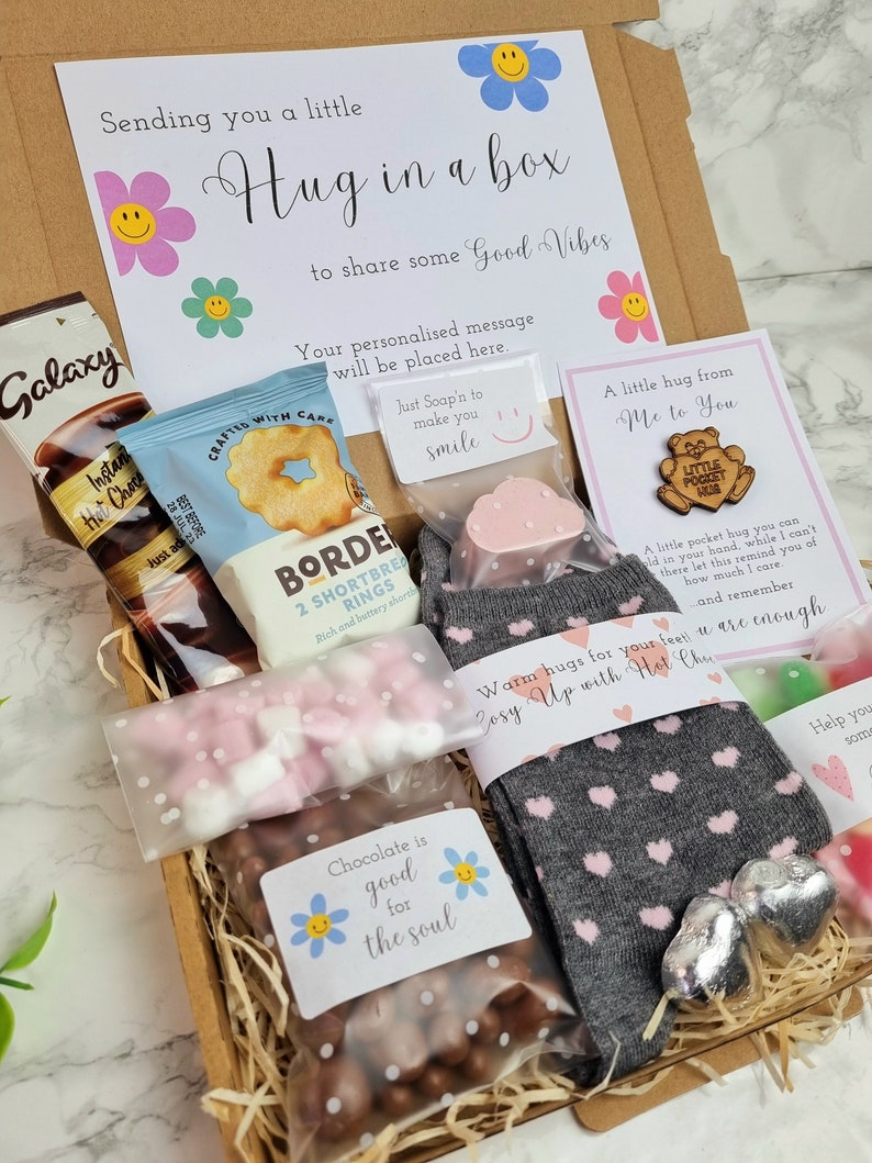May include: A gift box filled with various items, including chocolate, hot chocolate, socks, and a small wooden sign that says "A little hug from me to you". The box is decorated with a sign that says "Sending you a little hug in a box to share some good vibes".