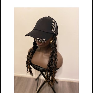 Pode incluir: Um boné de beisebol preto com três anéis metálicos prateados presos à aba e um detalhe de corrente prateada decorativa fixada por tachas na lateral. O boné é exibido em uma cabeça de manequim com cabelos longos, escuros e ondulados.