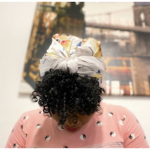 May include: A person with curly black hair is wearing a white satin headscarf with a yellow and blue floral print. The headscarf is tied in a knot at the top of their head. They are wearing a pink shirt with black and white yin yang symbols.