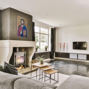 May include: A living room with a fireplace, a mosaic artwork, and a modern entertainment center. The room features a gray sofa, black curtains, and a set of wooden nesting tables. The floor is tiled in dark gray.