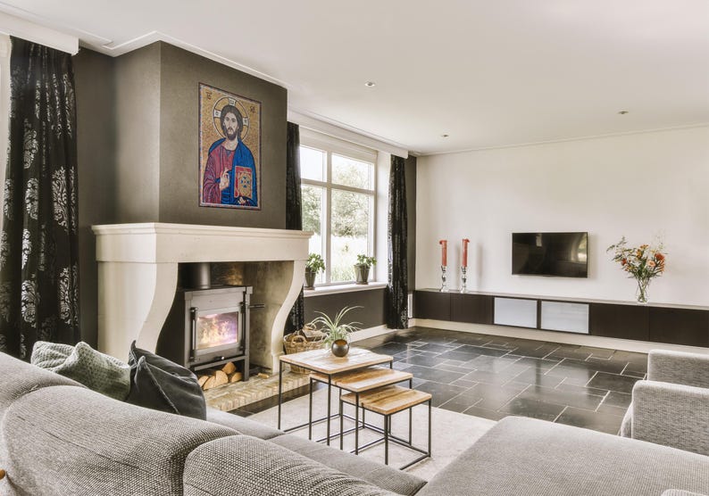 May include: A living room with a fireplace, a mosaic artwork, and a modern entertainment center. The room features a gray sofa, black curtains, and a set of wooden nesting tables. The floor is tiled in dark gray.