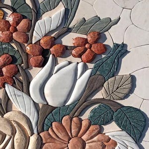 Op de afbeelding: Een bloemenmozaïek met een witte tulp, een perzikroze madeliefje en diverse andere bloemen en bladeren. Het mozaïek is gemaakt van steen en staat tegen een lichte achtergrond.