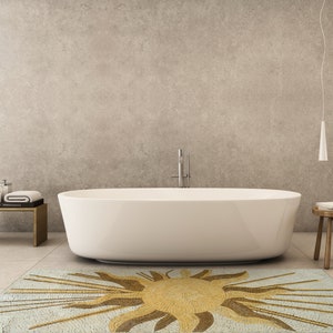 May include: A white bathtub sits on a light blue and gold mosaic rug with a sun design. The rug is in a modern bathroom with a concrete wall and a wooden stool.