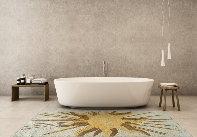 May include: A white bathtub sits on a light blue and gold mosaic rug with a sun design. The rug is in a modern bathroom with a concrete wall and a wooden stool.