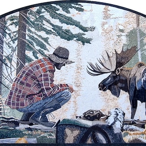 May include: Mosaic artwork depicting a man in a plaid shirt and hat squatting near a moose in a forest setting. The artwork features a detailed mosaic style with various shades of brown, green, and blue, creating a naturalistic scene. The moose has large antlers.