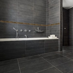 May include: A modern bathroom with dark grey tiled walls and flooring. A white bathtub is built into the wall, with chrome fixtures and a small shelf. A decorative border with gold accents runs along the wall.