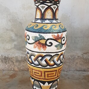 May include: A large, decorative vase made of mosaic tiles. The vase is decorated with a geometric pattern in shades of brown, yellow, and white. The vase is standing on a concrete floor.