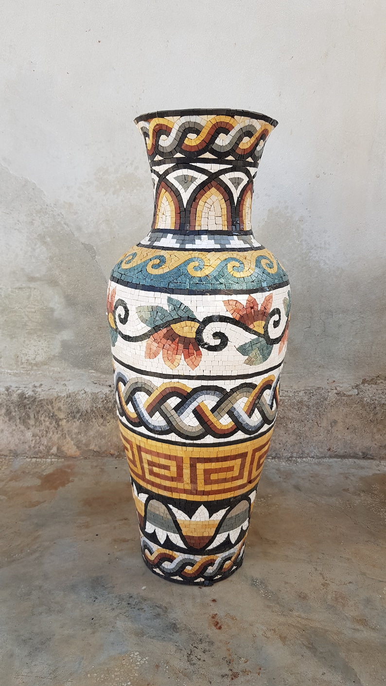 May include: A large, decorative vase made of mosaic tiles. The vase is decorated with a geometric pattern in shades of brown, yellow, and white. The vase is standing on a concrete floor.