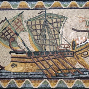 Op de afbeelding: Een mozaïekkunstwerk dat een schip met een zeil en roeispanen toont, tegen een achtergrond van blauwe en witte tegels. Het schip is omgeven door een decoratieve rand van gele en bruine tegels.