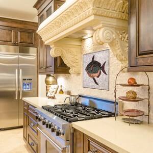 May include: A luxurious kitchen with dark wood cabinetry, a stainless steel refrigerator, and a gas range. A decorative mosaic of a fish hangs above the range. The kitchen features a light-colored countertop and a decorative range hood.