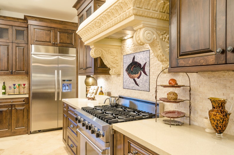 May include: A luxurious kitchen with dark wood cabinetry, a stainless steel refrigerator, and a gas range. A decorative mosaic of a fish hangs above the range. The kitchen features a light-colored countertop and a decorative range hood.