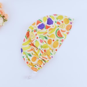 Puede incluir: Un gorro de quirófano de tela amarillo y blanco con un estampado de frutas coloridas. El gorro tiene un cierre de cordón.