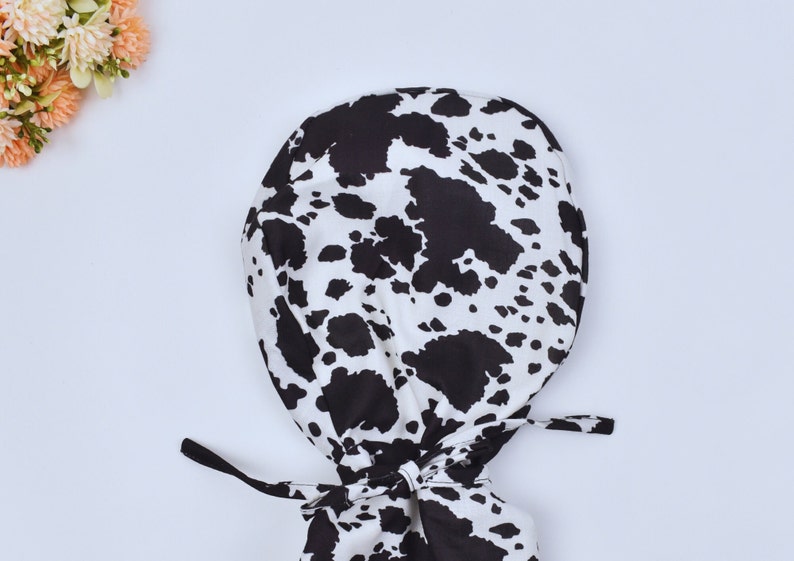 May include: A black and white cow print surgical scrub hat with ties.